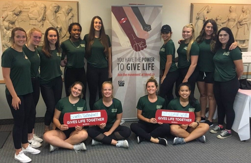 Womens volleyball players posing with give blood display