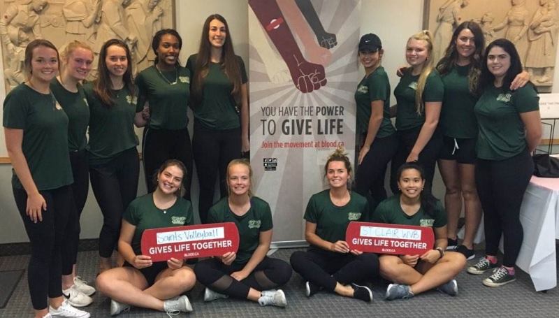 Womens volleyball players posing with give blood display