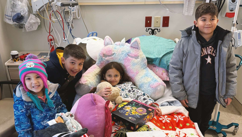 Lainey McMahon sits in a hospital bed surrounded by loved ones